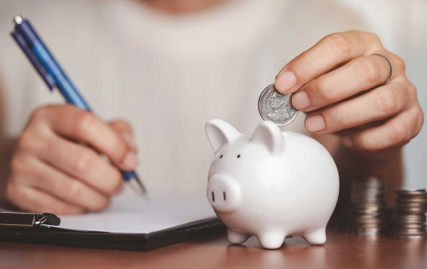 Sparschwein auf dem Tisch. Person mit Stift und Geld in der Hand, die Geld in das Schwein wirft