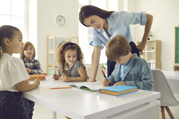 Kinder sitzen an einem Tisch im Klassenraum und machen Schulaufgaben. Lehrerin steht hinten den Kindern.